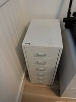 White metal storage cabinet from a top angle showing six drawers with silver handles and surface marks.
