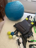 Photo showing blue deflated exercise ball, three tennis or therapy balls, black and white strap, and Mustang Survival Khimera flotation vest on bed.