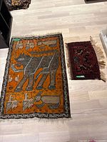 View of both Persian rugs laid flat on light wood floor showing overall size and design; larger orange rug with animals and smaller red rug with geometric motifs