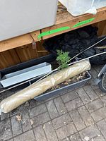 Full view of three plastic planter boxes, rolled up gardening fabric, and bamboo sticks placed on the ground under a wooden shelf.