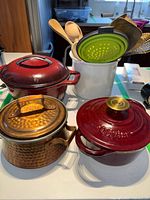 All pots and kitchen utensils arranged on a table, showing medium red cast iron pot, small copper-colored pot, and wooden utensils in white container