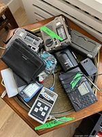 Photo showing multiple radios, tape recorder, thermometer, battery charger, hand-held devices, and accessories on a metal tray.