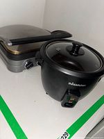 Photo of Thinkkitchen rice cooker with glass lid and Cuisinart Griddler appliance together on a white surface showing front switch and dial control.
