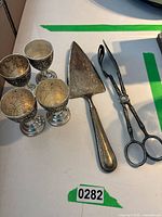 Overall shot showing four vintage grapevine engraved sterling silver wine goblets, a pair of sterling silver tongs, and a sterling silver cake server on a white surface.