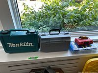 Photo showing three tool boxes/cases on a window sill: a teal Makita case, a grey DeWalt case, and two smaller plastic cases stacked with a retractable blade on top.
