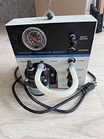 Front view of Vacuum/Pressure Station showing vacuum gauge, control knobs, tubing, and power cord on a wooden surface.