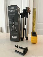 Tripod folded beside box, smartphone clamp and tape measure for scale