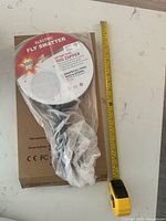 Swatter in plastic on top of retail box with tape measure for scale