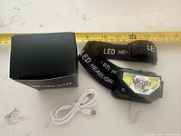 Headlamp, strap, charging cable and box laid out with measuring tape