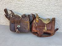 Shows two vintage leather saddle purses side by side on concrete against a textured wall.