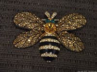 Front view of large gold-tone bee brooch adorned with amber rhinestones on wings, a central large amber rhinestone, clear rhinestone stripes on the black enamel abdomen, and two green rhinestones for eyes.