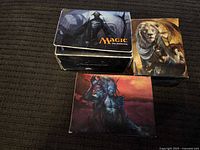 Photo showing three Magic the Gathering card deck boxes arranged on dark fabric. Boxes have distinct character artwork on lids.
