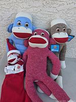 Four sock monkey plush toys displayed against a textured wall, including blue, red, brown monkeys and a smaller santa-themed stocking monkey with tags.