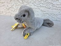 Full view of soft gray plush seal lying on surface with visible tags on ear and around neck