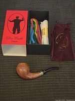 Photo showing the Don Carlos tobacco pipe, original box, velvet pouch, and cleaning accessories laid out on a textured dark surface.