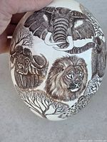 Close-up of an etched ostrich egg showing African wildlife including elephant, lion, buffalo, and trees.