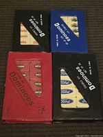 Four domino sets shown from above in original cardboard cases with visible company logos on the domino tiles.