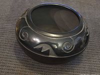 Large black pottery bowl signed Santa Clara Pueblo with incised geometric and spiral motifs, polished surface visible on a fabric background.