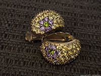 Close-up photo showing a pair of sterling silver clip-on earrings encrusted with yellow, purple, and green rhinestones arranged in floral patterns.