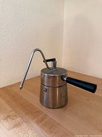 Photo showing vintage stove top espresso maker with stainless steel body and long angled spout, black plastic handle, placed on wooden surface beside light-colored wall.