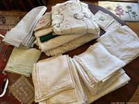 Wide view of the stacked assortment of antique linens including tablecloths, napkins, and serving cloths in various sizes and white to cream colors.