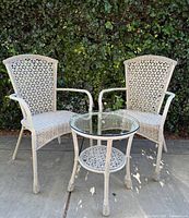 Front view of two sand woven wicker patio chairs flanking a round wicker table with a clear glass top and patterned lower shelf, set outdoors on a concrete surface with greenery backdrop.