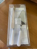 Clear plastic packaging showing the dessert server set with white handles and stainless steel blades on a wooden surface