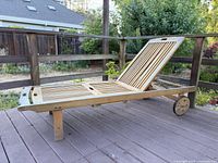 Side view of teak chaise lounge chair on outdoor deck, showing slatted adjustable backrest and wheels at one end.