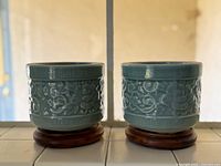 Front view showing two teal glazed porcelain vases with embossed floral designs on wooden bases.