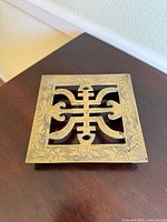 Square brass trivet with central Asian-style cutout and floral etched border, viewed from above