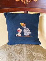 Front view of the needlepoint pillow showing a lady in a white dress and bonnet interacting with a small dog on a blue background.