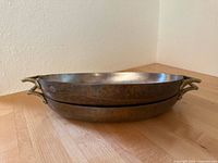 Two stacked large oval copper au gratin pans with brass handles, showing their aged copper exterior.