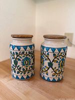 Pair of vintage large ceramic kitchen jars with hand-painted leaf and flower designs, showing geometric blue triangle borders and wood lids.