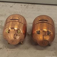 Front view of 2 copper piggy banks showing facial details, coin slots, and feet
