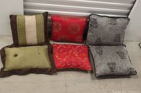 Photo showing six cushions: two grey with prints, two red with gold patterns, one green-beige-brown striped, and one rectangular green with button and brown border