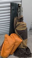 Multiple rolled and folded tent fabric pieces in various colors including orange, brown, and black, along with bundled metal poles in a greenish bag.