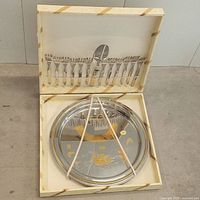 Open box showing round metal serving platter with gold-tone decoration secured with straps, and matching cutlery set lined in lid.