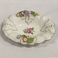 Front angled view of white porcelain dish showing hand painted floral motifs, pink rose center, multicolor flowers, and gold leaf outlines on leaves.