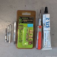 Photo showing Greenlee combination drill/tap bit in packaging, three loose hand taps, screwdriver, and lubricant tube laid out on surface.