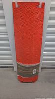 Heavy duty orange cord cover mat standing upright against a white wall, showing tread plate pattern and top cutouts for cords