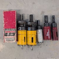 Photo showing seven hole saws including two large yellow, one white, two red, black hex shank drill adapter, and red box with label.