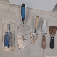Five masonry tools laid out on the floor showing handles and blades with cement residue.