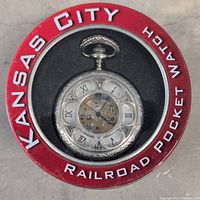 Top view of the pocket watch inside factory metal tin with red lid labeled 'Kansas City Railroad Pocket Watch'. Visible gears through watch face.