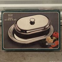 Box front with image of the Athenas by Tramontina stainless steel covered butter dish showing the oval shape, polished finish, and knob handle on lid.