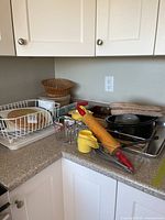 Photo of kitchen corner counter displaying two glass measuring jugs, yellow plastic citrus juicer, wooden rolling pin with red handles, baking trays, roasting pan, oven mitts, dish rack with plastic base, and small kitchen items.