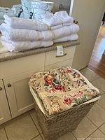 Stack of 4 white bath towels and 4 facecloths on kitchen counter alongside beige floral laundry hamper with lace trim.