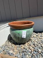 Full view of a large green ceramic pot with a terracotta rim sitting on gravel outdoors.
