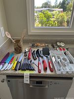 Full view of cutlery and kitchen utensils arranged on countertop showing colorful knives, black handled knives and forks, wooden spoons in ceramic cup, spatulas, graters, and serving spoons.