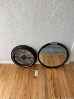 Both black plastic framed wall clock and round mirror displayed on hardwood floor against textured white wall.