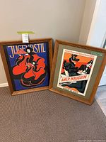 Two wood framed posters leaning against a wall on carpet. One poster has 'Jugendstil' text and a stylized woman in red and black. The other poster advertises a theatre show in Liège with a man and a woman. Both have oak wood frames.